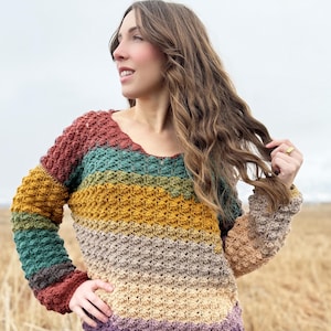 May include: A multi-coloured crochet sweater with a striped pattern in shades of rust, teal, mustard yellow, beige, and purple. The sweater has a v-neck and long sleeves.