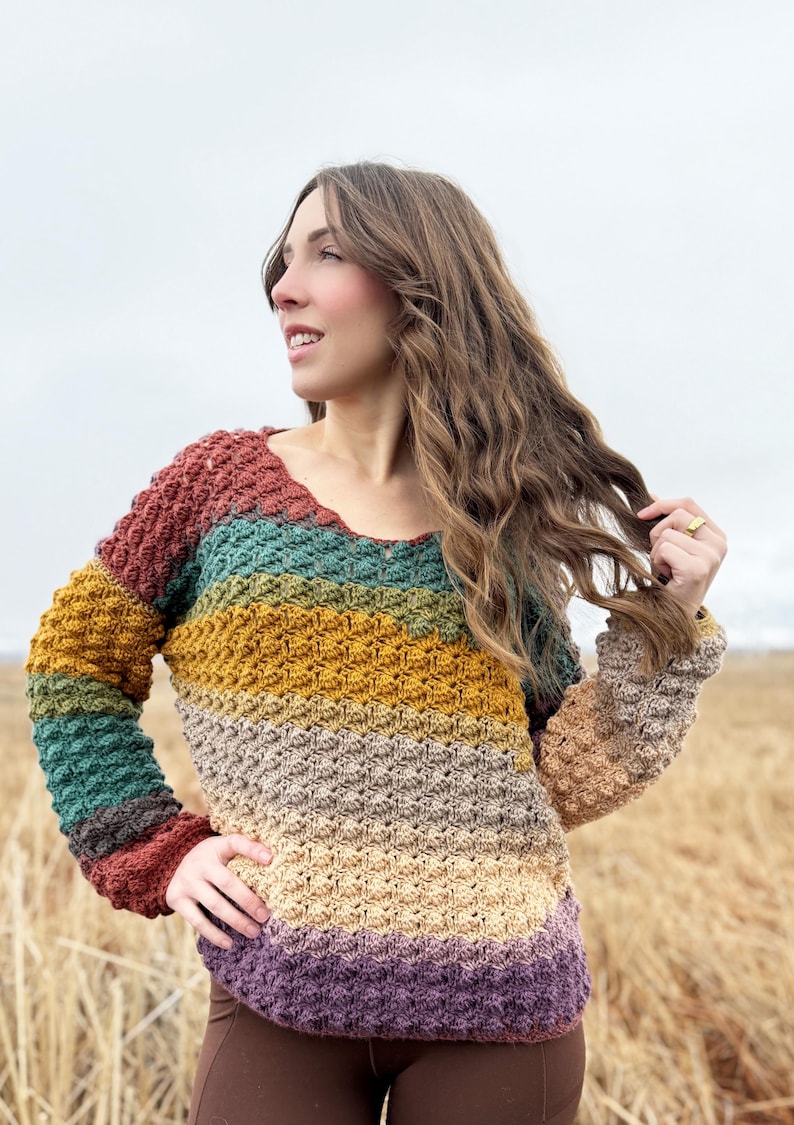 May include: A multi-coloured crochet sweater with a striped pattern in shades of rust, teal, mustard yellow, beige, and purple. The sweater has a v-neck and long sleeves.