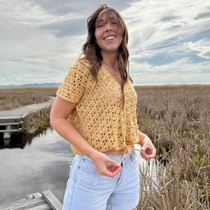 May include: A mustard yellow crochet top with a V-neck and short sleeves. The top is paired with light blue denim shorts. The outfit is worn outdoors with a cloudy sky and marshy background.