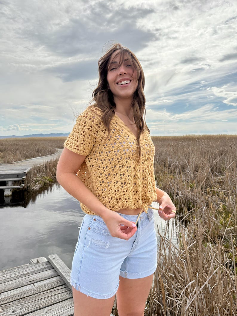 May include: A mustard yellow crochet top with a V-neck and short sleeves. The top is paired with light blue denim shorts. The outfit is worn outdoors with a cloudy sky and marshy background.