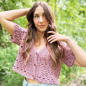 May include: A short-sleeved, dusty rose crochet cardigan with a V-neck and button closure. The open weave design reveals a glimpse of the wearer's skin. The cardigan is paired with light blue denim shorts. The background is a blurred green.