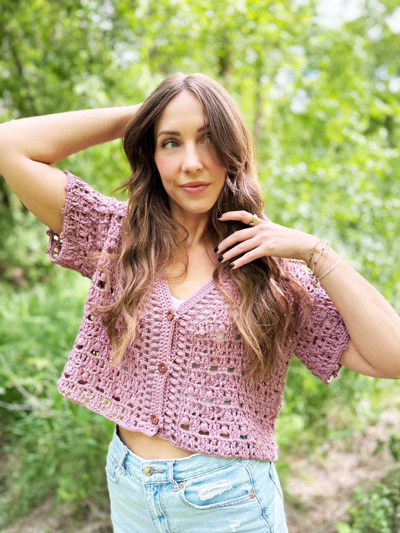 May include: A short-sleeved, dusty rose crochet cardigan with a V-neck and button closure. The open weave design reveals a glimpse of the wearer's skin. The cardigan is paired with light blue denim shorts. The background is a blurred green.