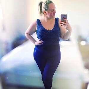 May include: A woman wearing a blue tank top and dark blue leggings. She is standing in front of a white wall and holding a phone in her hand.