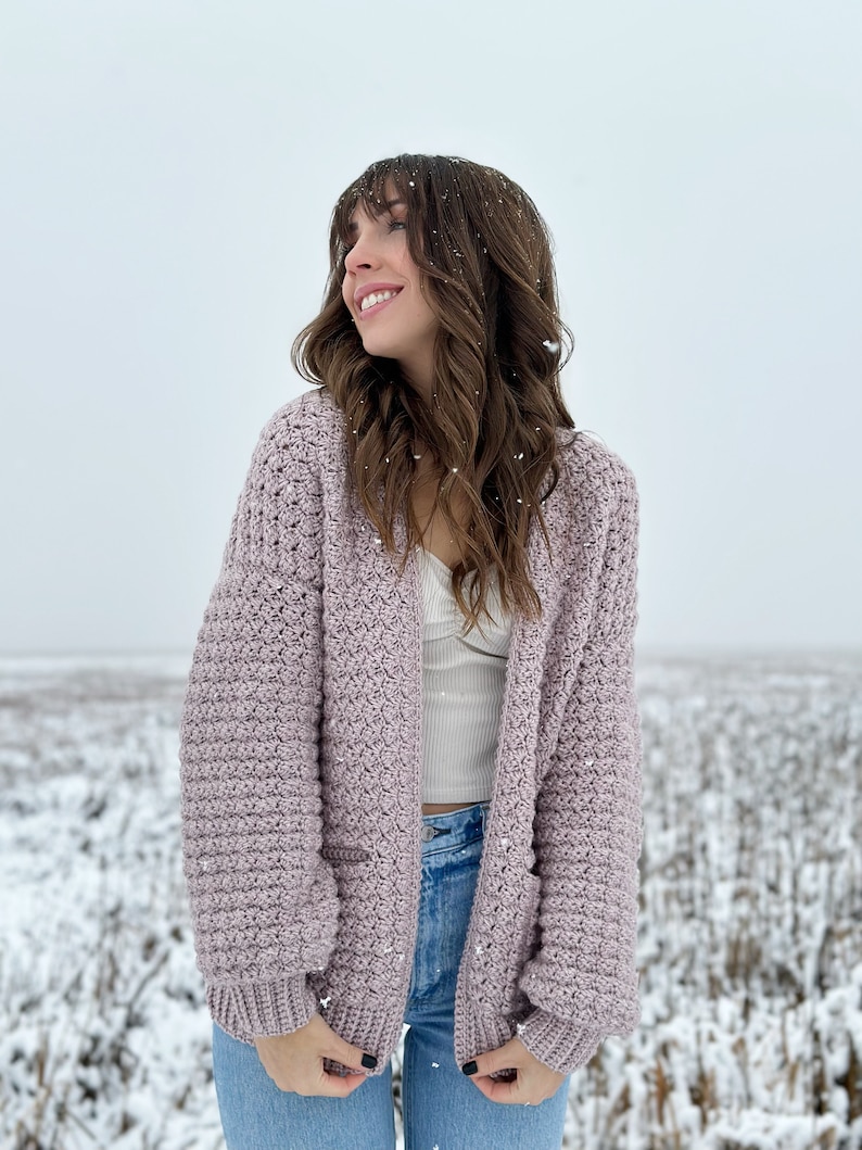 May include: A woman wearing a light purple crocheted cardigan with pockets in a snowy field.