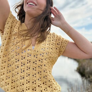 May include: A yellow crochet top with a V-neck and short sleeves. The top has a drawstring at the waist and a detailed openwork pattern. The top is paired with light blue denim shorts. The background features a body of water and a cloudy sky.
