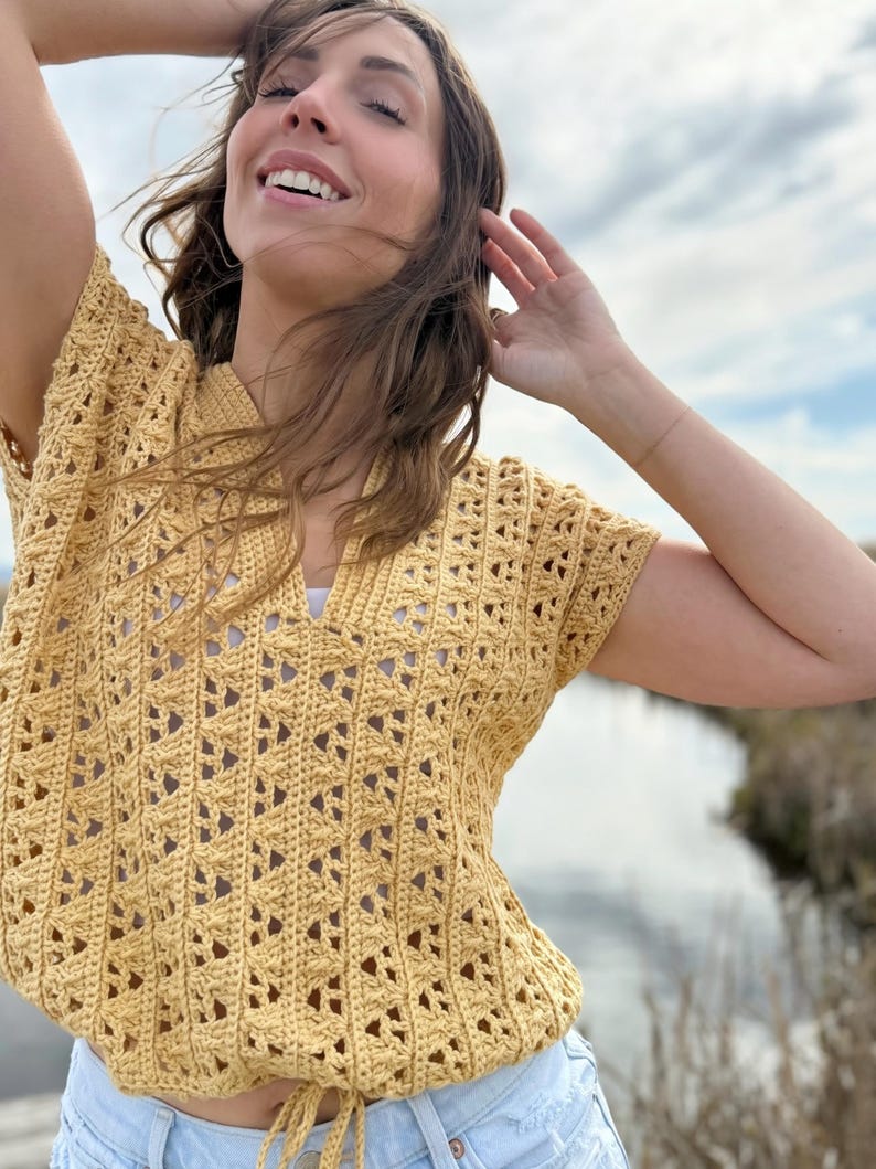 May include: A yellow crochet top with a V-neck and short sleeves. The top has a drawstring at the waist and a detailed openwork pattern. The top is paired with light blue denim shorts. The background features a body of water and a cloudy sky.