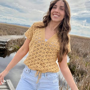 May include: Mustard yellow crochet top with a V-neck and short sleeves. The top has a drawstring waist and a geometric openwork pattern. The model is wearing light blue denim shorts. The background includes a wooden walkway, water, and tall grass.