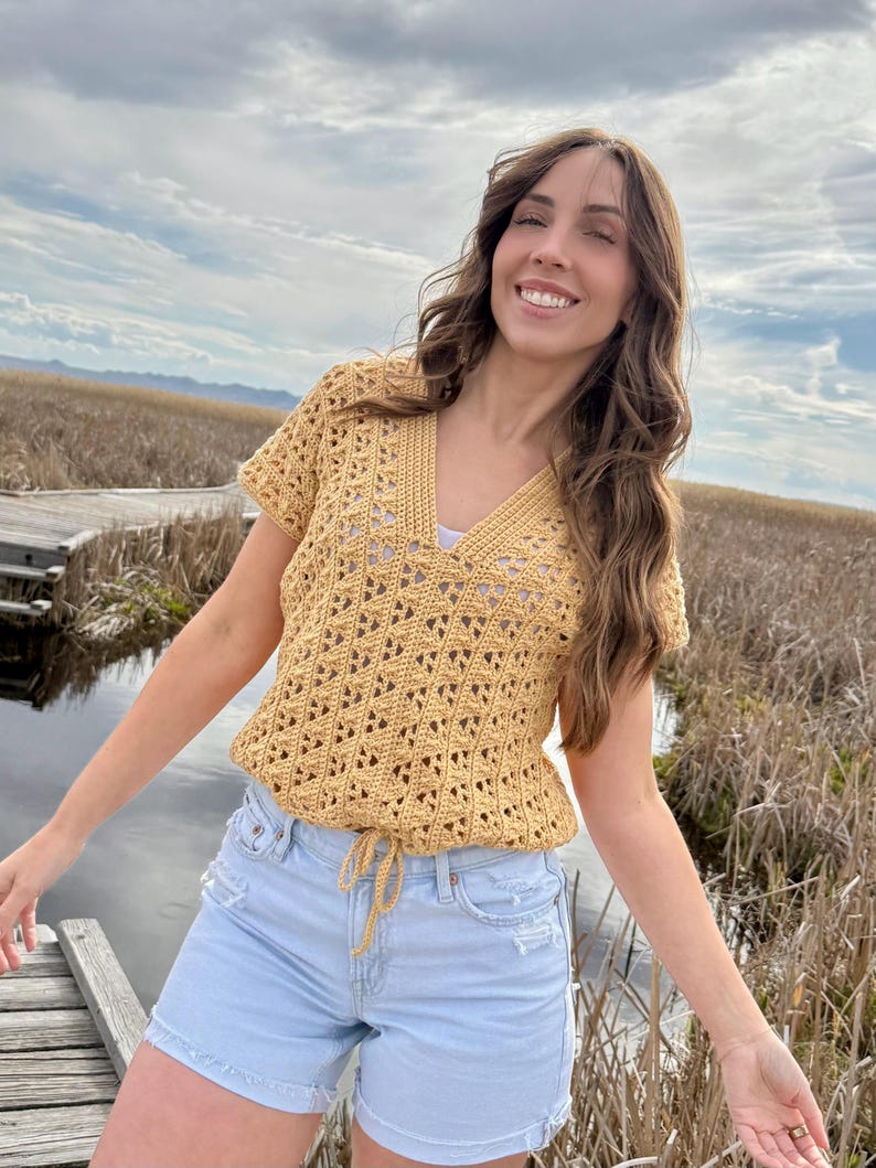 May include: Mustard yellow crochet top with a V-neck and short sleeves. The top has a drawstring waist and a geometric openwork pattern. The model is wearing light blue denim shorts. The background includes a wooden walkway, water, and tall grass.