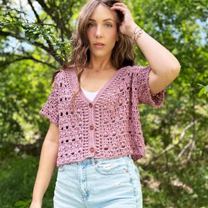 May include: A dusty rose crochet top with short sleeves and a button-up front. The top is paired with light blue denim shorts. The background features green foliage and a blue sky.