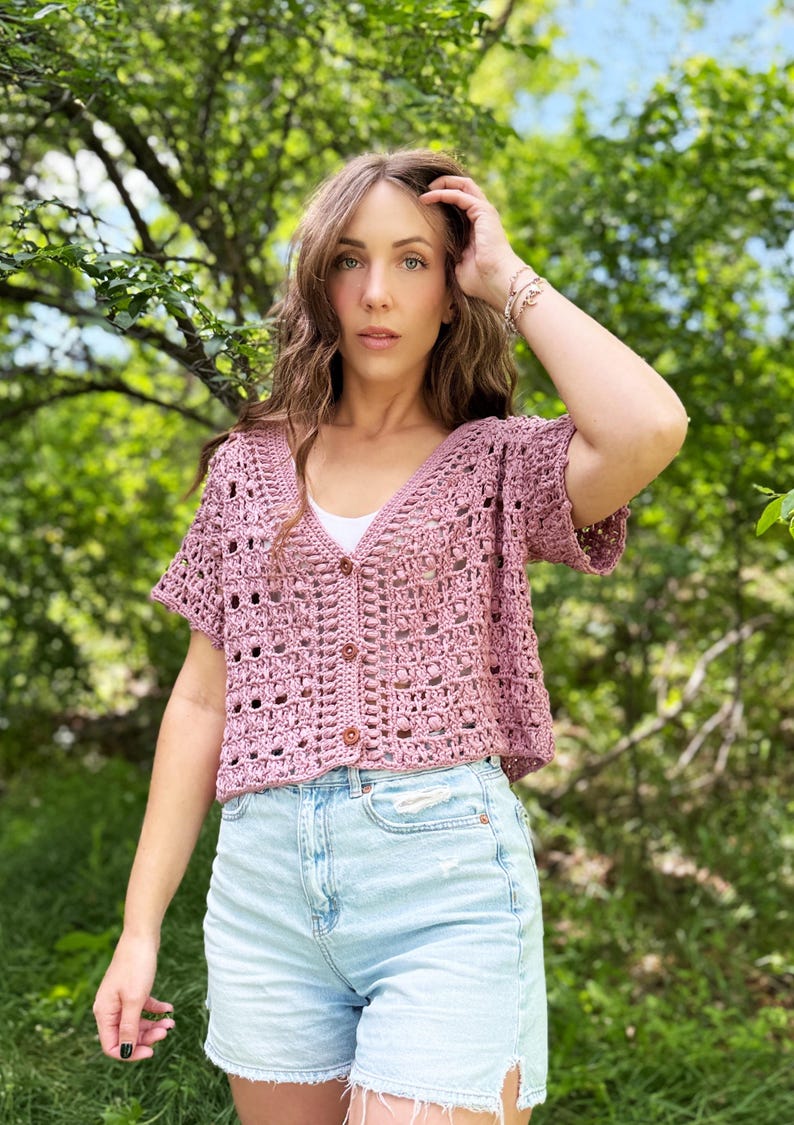 May include: A dusty rose crochet top with short sleeves and a button-up front. The top is paired with light blue denim shorts. The background features green foliage and a blue sky.