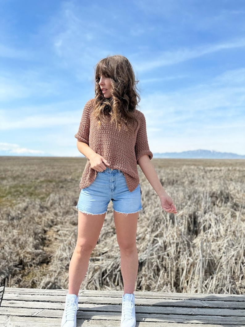 Puede incluir: Una mujer con un su&eacute;ter de punto marr&oacute;n, pantalones cortos de mezclilla azul claro y zapatillas blancas. Est&aacute; de pie en una plataforma de madera con un campo de hierba seca en el fondo.