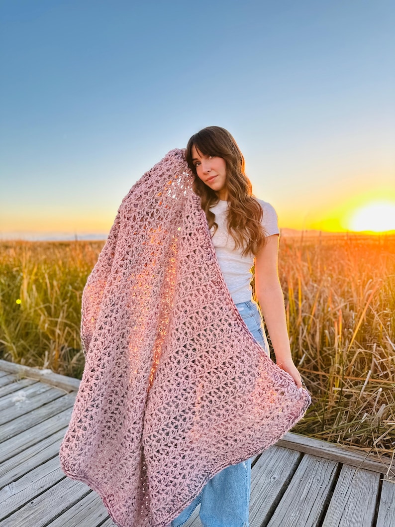 May include: A woman wearing a white shirt and blue jeans is holding a large, crocheted blanket in a dusty rose color. The blanket has a delicate, openwork pattern and is draped over her shoulder. The background is a field of tall grass with a sunset in the distance.
