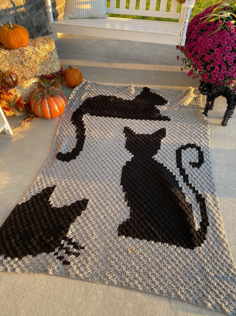 Op de afbeelding: Een grijs gebreid deken met zwarte katten silhouetten. Het deken ligt op een betonnen veranda met pompoenen en hooibalen op de achtergrond.