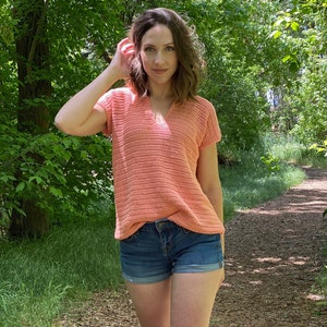 以下が含まれることがあります： 女性は、短い袖とVネックのコーラル色のクロシェトップを着ています。彼女はデニムのショートパンツを履いており、森の中の小道に立っています。
