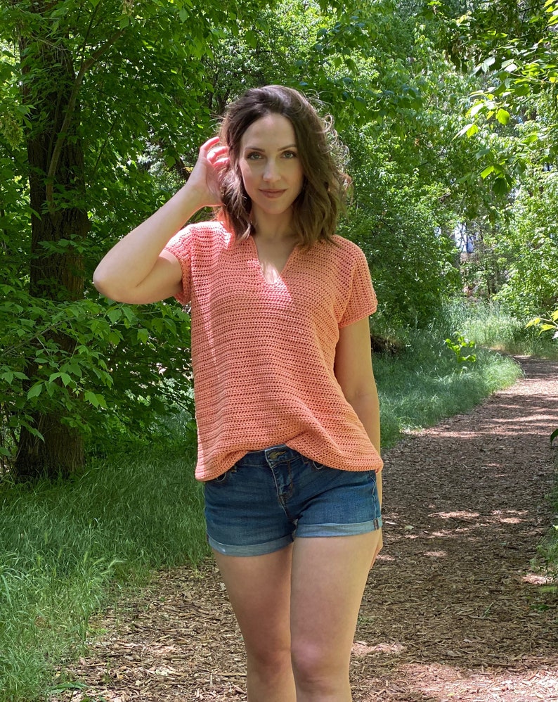以下が含まれることがあります： 女性は、短い袖とVネックのコーラル色のクロシェトップを着ています。彼女はデニムのショートパンツを履いており、森の中の小道に立っています。