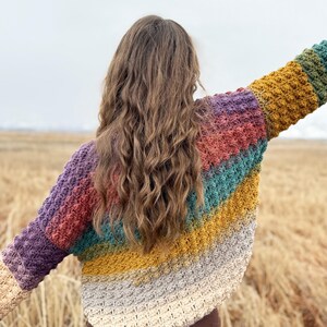 May include: A multi-coloured crochet sweater with a striped pattern in shades of purple, red, teal, yellow, and grey. The sweater is loose-fitting and has a relaxed silhouette.