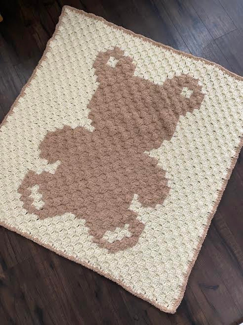 Pode incluir: Uma manta de croch&ecirc; branca com uma silhueta de urso de croch&ecirc; marrom no centro.