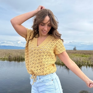 May include: A yellow crochet top with a V-neck and short sleeves. The top has a drawstring at the waist and is paired with light blue denim shorts. The outfit is worn outdoors near a body of water.
