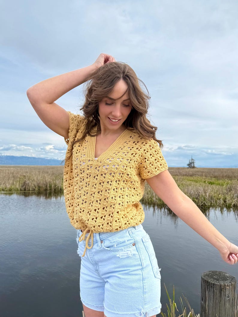 May include: A yellow crochet top with a V-neck and short sleeves. The top has a drawstring at the waist and is paired with light blue denim shorts. The outfit is worn outdoors near a body of water.