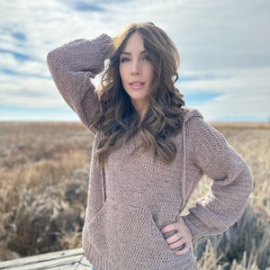May include: A mauve knitted hoodie with a large pocket and a white trim around the bottom. The hoodie is worn by a person with long brown hair, standing in a field.