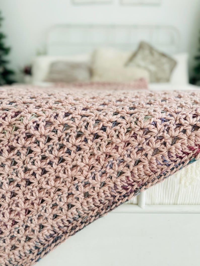 May include: A soft, pink crocheted throw blanket with a subtle multi-colored pattern. The blanket is draped over a white bed with pillows.