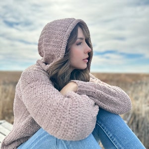 May include: A woman wearing a mauve knitted hooded sweater and blue jeans sits with her arms crossed, looking off to the side. The background is a blurred out field.