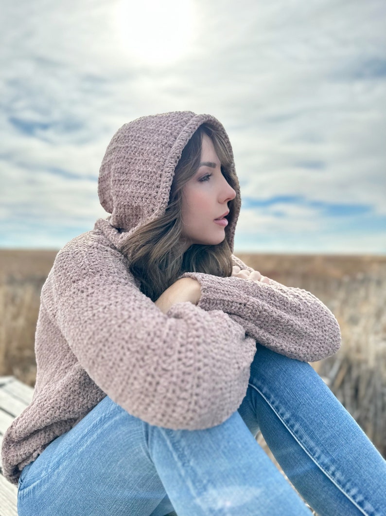 May include: A woman wearing a mauve knitted hooded sweater and blue jeans sits with her arms crossed, looking off to the side. The background is a blurred out field.