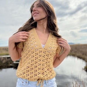 May include: Mustard yellow crochet top with a V-neck and short sleeves. The top features an open, geometric pattern and a drawstring at the waist. The model is wearing light blue denim shorts.