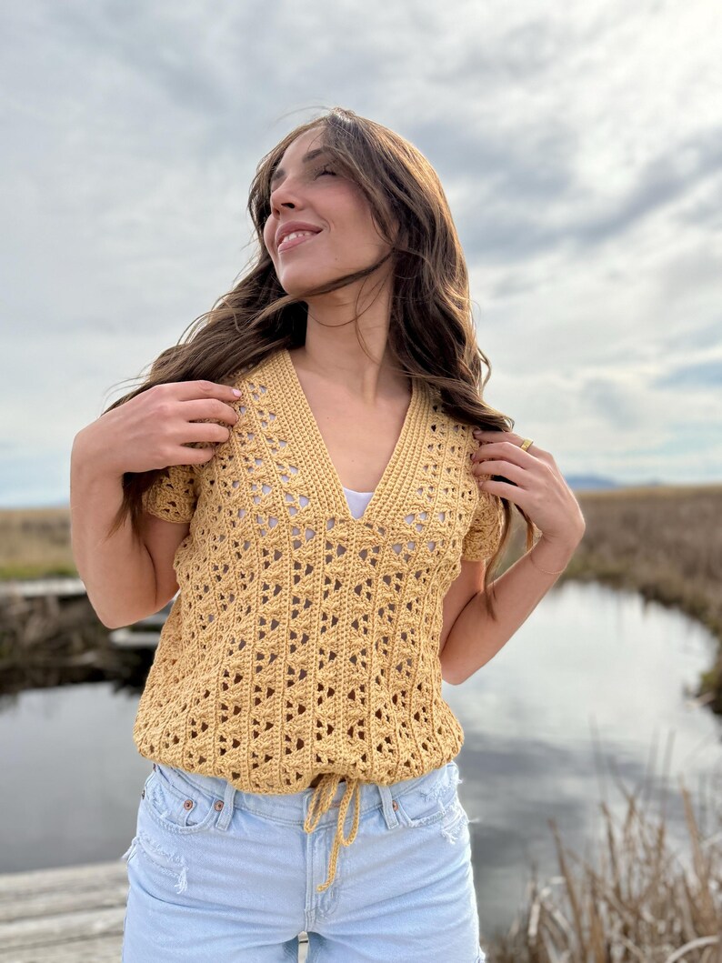 May include: Mustard yellow crochet top with a V-neck and short sleeves. The top features an open, geometric pattern and a drawstring at the waist. The model is wearing light blue denim shorts.