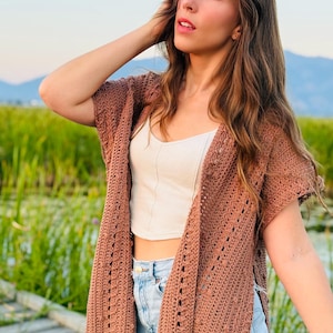 May include: A brown crochet cardigan with short sleeves and an open front design, worn over a white sleeveless crop top and light blue denim shorts. The cardigan features a textured, open-knit pattern with vertical detailing along the front edges.