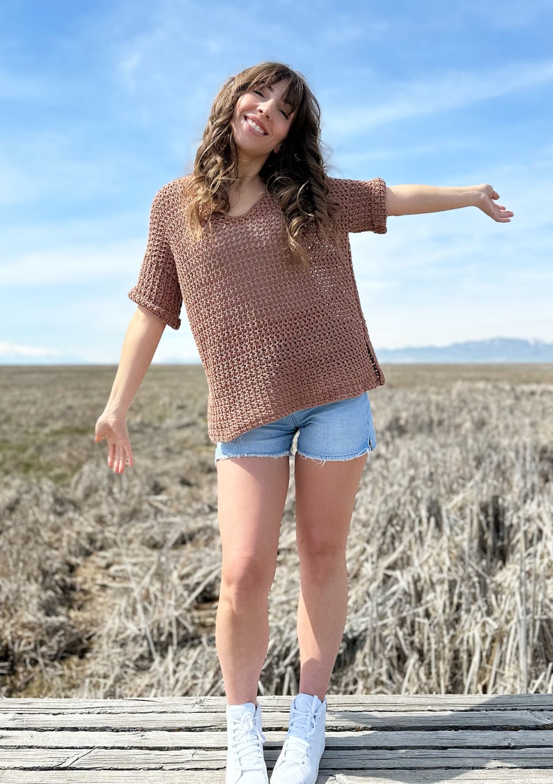 Puede incluir: Una mujer con un top de crochet marr&oacute;n de manga corta, pantalones cortos de mezclilla y zapatillas blancas de ca&ntilde;a alta. Est&aacute; de pie en una plataforma de madera con un campo de hierba seca en el fondo.