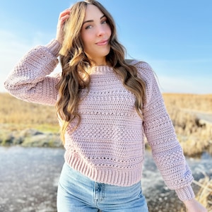 May include: A light pink crochet sweater with long sleeves and a round neckline. The sweater has a textured stitch pattern and is paired with blue jeans. The background features a field and a clear blue sky.