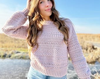 May include: A light pink crochet sweater with long sleeves and a round neckline. The sweater has a textured stitch pattern and is paired with blue jeans. The background features a field and a clear blue sky.