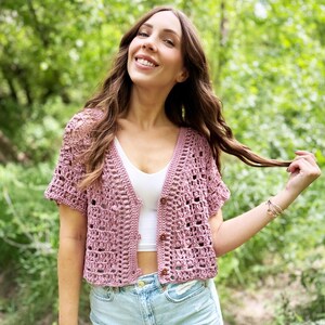 May include: A short-sleeved, open-weave, dusty rose crochet cardigan with wooden buttons. The cardigan is worn over a white top and paired with light blue denim shorts. The background is a blurred green forest.