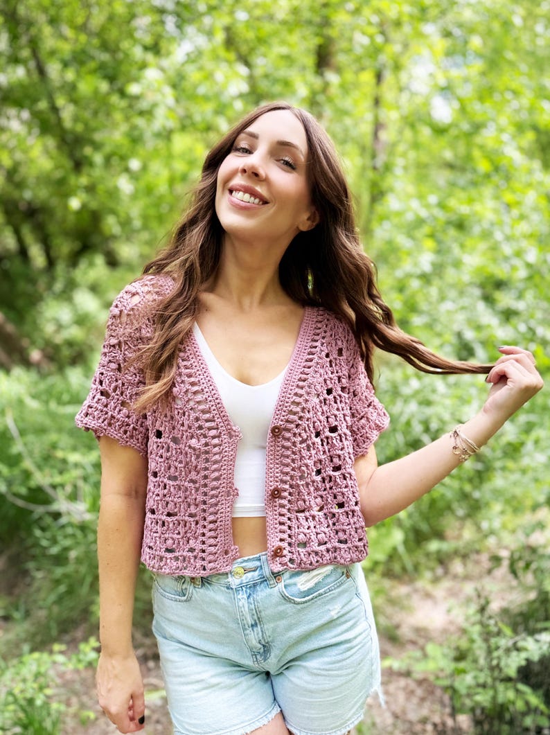 May include: A short-sleeved, open-weave, dusty rose crochet cardigan with wooden buttons. The cardigan is worn over a white top and paired with light blue denim shorts. The background is a blurred green forest.