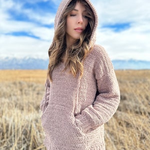 May include: A soft, light pink crocheted hooded sweater with a large pocket on the front. The sweater is worn by a person standing in a field of dry grass.