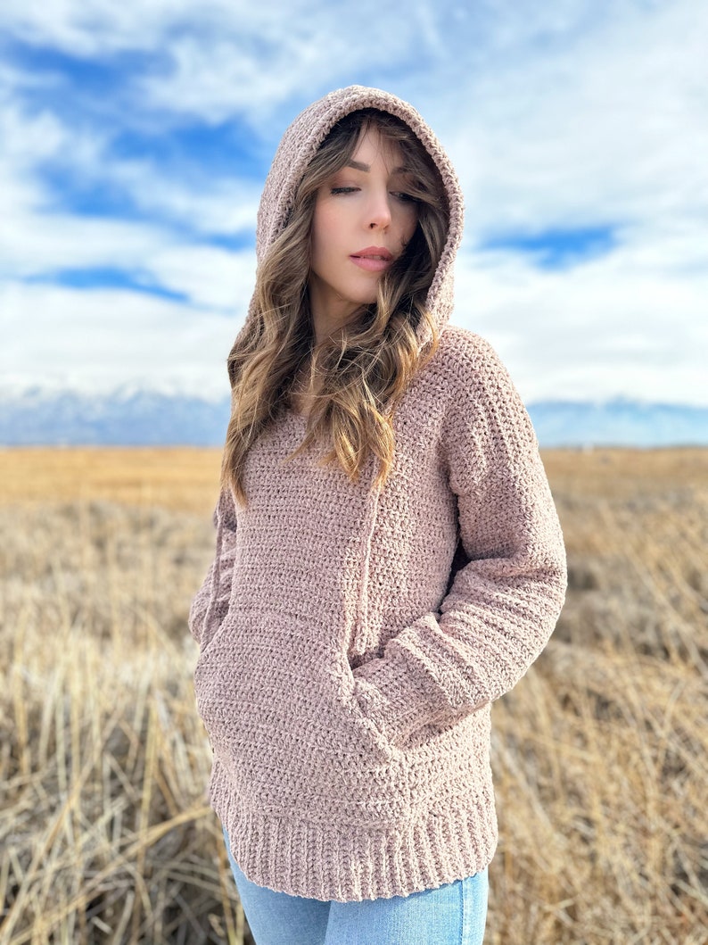 May include: A soft, light pink crocheted hooded sweater with a large pocket on the front. The sweater is worn by a person standing in a field of dry grass.