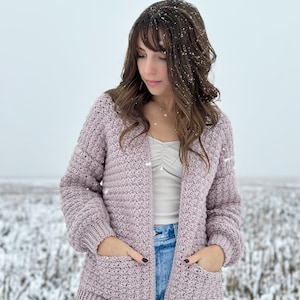 May include: A woman wearing a light purple crocheted cardigan with pockets and long sleeves. She is standing in a snowy field.