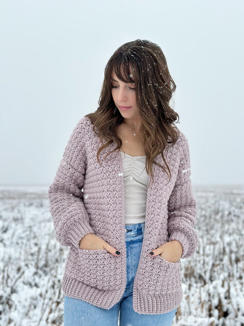 May include: A woman wearing a light purple crocheted cardigan with pockets and long sleeves. She is standing in a snowy field.