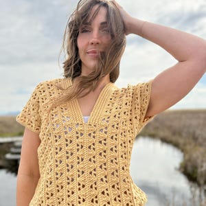 May include: A yellow crochet top with a V-neck and short sleeves. The top has a drawstring at the waist and a geometric openwork pattern. The top is made of a soft, textured yarn.