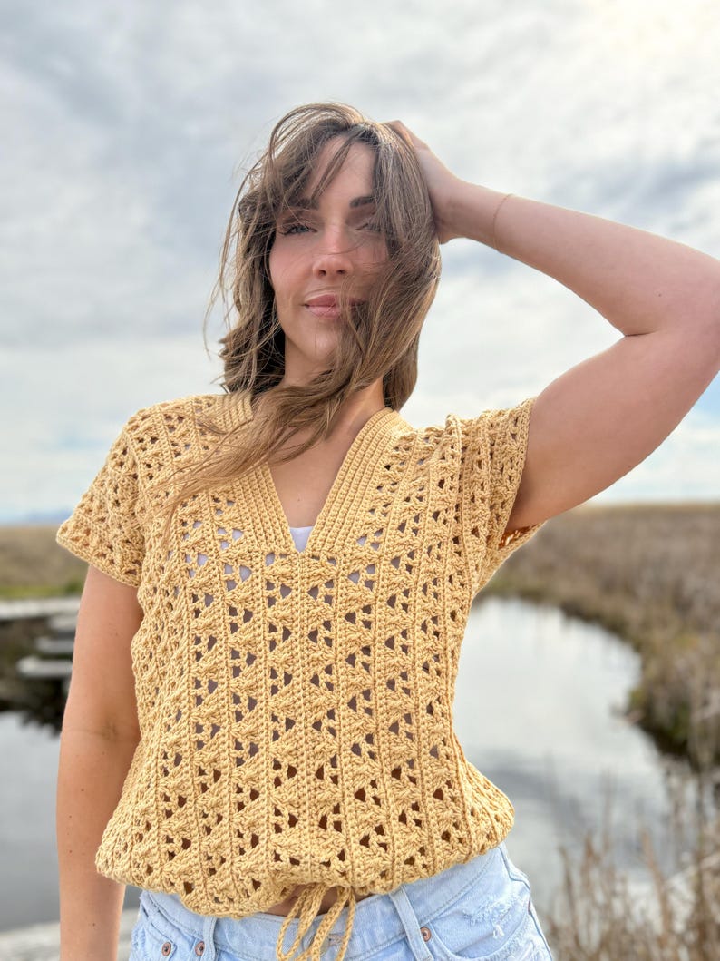 May include: A yellow crochet top with a V-neck and short sleeves. The top has a drawstring at the waist and a geometric openwork pattern. The top is made of a soft, textured yarn.