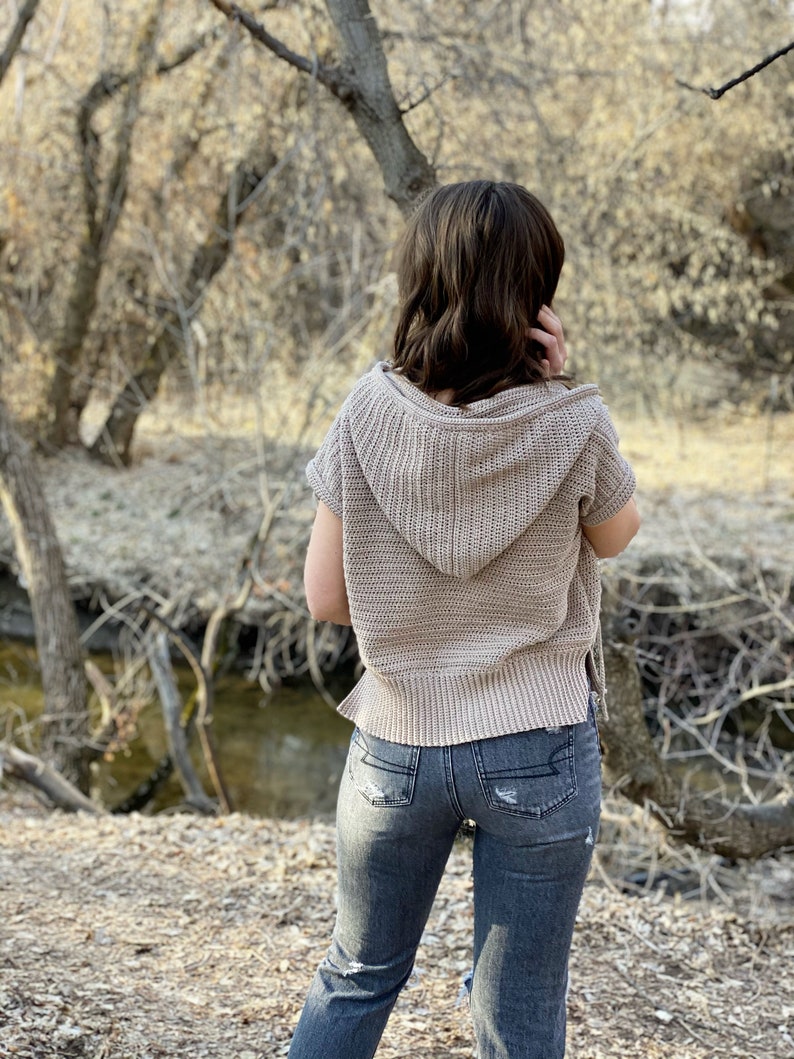 May include: A woman wearing a light brown hooded sweater with short sleeves and distressed blue jeans. She is standing in a wooded area with a small stream in the background.