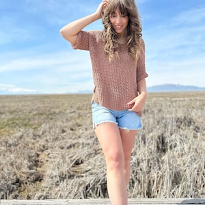 Puede incluir: Una mujer con un top de crochet marr&oacute;n y pantalones cortos de mezclilla se para en una plataforma de madera. El fondo es un campo de hierba seca y un cielo azul.