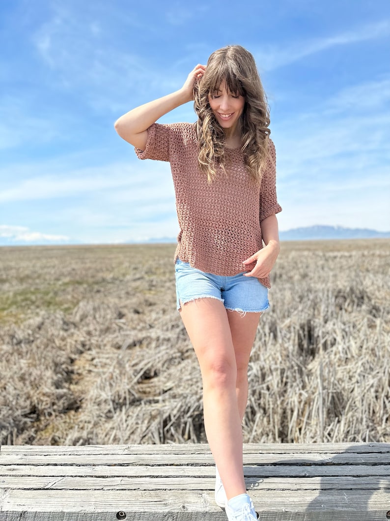 Puede incluir: Una mujer con un top de crochet marr&oacute;n y pantalones cortos de mezclilla se para en una plataforma de madera. El fondo es un campo de hierba seca y un cielo azul.