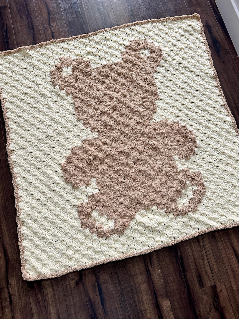 Pode incluir: Um cobertor de beb&eacute; de croch&ecirc; com um desenho de urso castanho. O cobertor &eacute; feito com um ponto texturizado e tem um fundo branco com um urso castanho no centro.