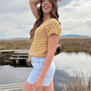 May include: A mustard yellow crochet top with short sleeves and a V-neck. The top is paired with light blue denim shorts. The outfit is worn outdoors near a body of water and a wooden dock.