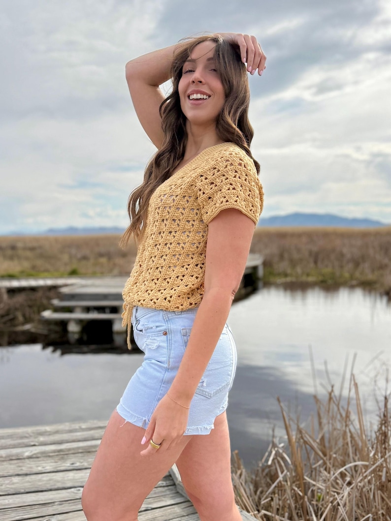 May include: A mustard yellow crochet top with short sleeves and a V-neck. The top is paired with light blue denim shorts. The outfit is worn outdoors near a body of water and a wooden dock.