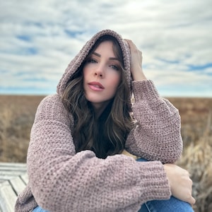 May include: A woman wearing a mauve hooded sweater with a soft, textured knit. She is sitting on a wooden bench with her legs crossed and her hand resting on her knee. The background is a field of tall grass.