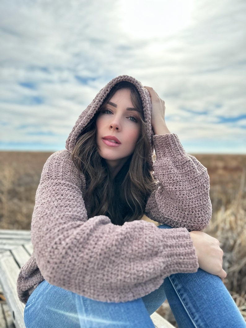 May include: A woman wearing a mauve hooded sweater with a soft, textured knit. She is sitting on a wooden bench with her legs crossed and her hand resting on her knee. The background is a field of tall grass.