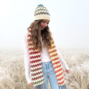 Puede incluir: Una mujer lleva un gorro blanco de ganchillo con un pompón de colores y una larga bufanda de ganchillo a rayas con flecos. La bufanda está hecha en tonos de rojo, naranja, amarillo, verde y marrón. Lleva una camisa blanca de manga larga y unos vaqueros azules.